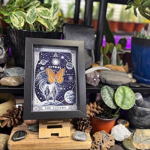 May include: A framed print of "The Lovers" tarot card. The artwork features a celestial background with a large orange butterfly, two figures holding hands, and the text "THE LOVERS". The frame is black.