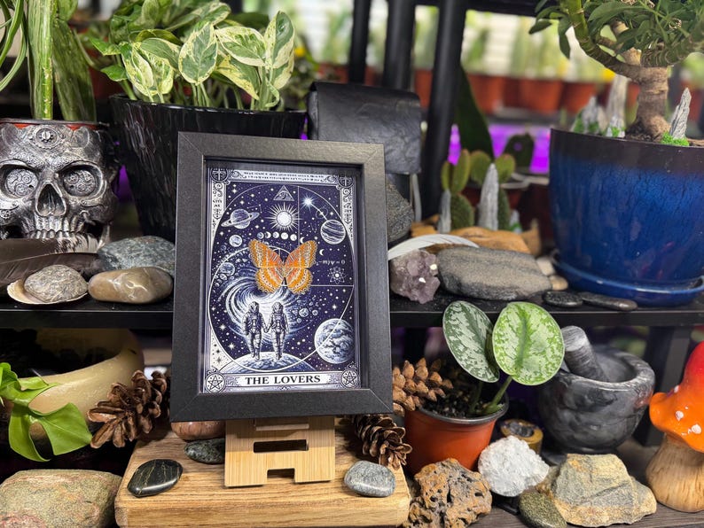 May include: A framed print of "The Lovers" tarot card. The artwork features a celestial background with a large orange butterfly, two figures holding hands, and the text "THE LOVERS". The frame is black.