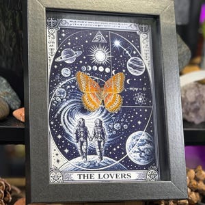 May include: Framed artwork with a celestial design, including planets, stars, and astronauts holding hands. An orange butterfly is centered on the image. The text "THE LOVERS" is at the bottom. The frame is dark grey.