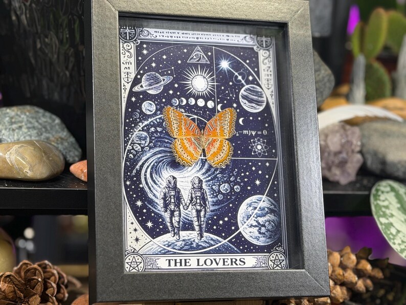 May include: Framed artwork with a celestial design, including planets, stars, and astronauts holding hands. An orange butterfly is centered on the image. The text "THE LOVERS" is at the bottom. The frame is dark grey.