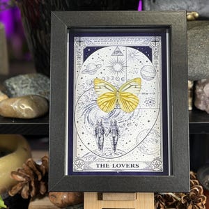 K&ouml;nnte beinhalten: Ein gerahmter Druck der Tarotkarte "Die Liebenden". Das Kunstwerk zeigt einen gelben Schmetterling, zwei Figuren in Raumanz&uuml;gen und Himmelselemente. Der Rahmen ist schwarz und der Druck hat ein blaues und wei&szlig;es Farbschema.
