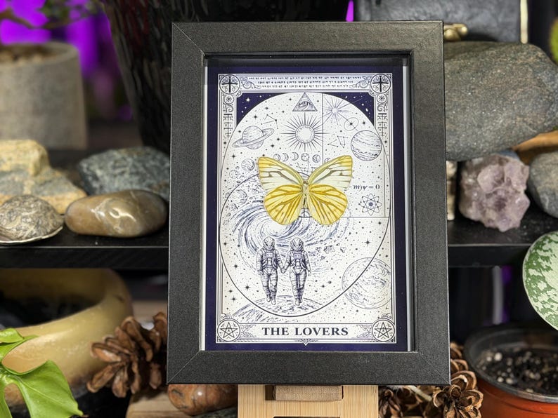 K&ouml;nnte beinhalten: Ein gerahmter Druck der Tarotkarte "Die Liebenden". Das Kunstwerk zeigt einen gelben Schmetterling, zwei Figuren in Raumanz&uuml;gen und Himmelselemente. Der Rahmen ist schwarz und der Druck hat ein blaues und wei&szlig;es Farbschema.