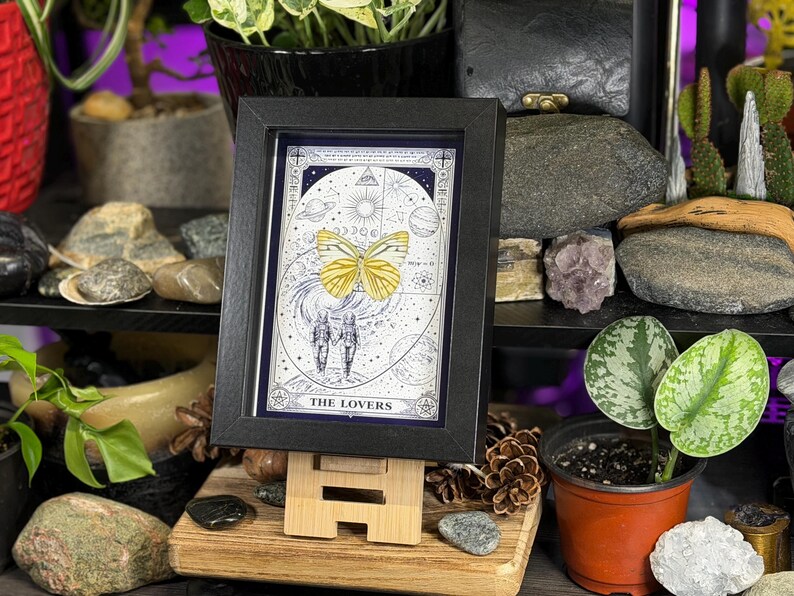 K&ouml;nnte beinhalten: Ein gerahmter Druck der Tarotkarte "Die Liebenden" mit einem gelben Schmetterling und zwei Figuren vor einem himmlischen Hintergrund. Der schwarze Rahmen wird auf einer Holzstaffelei pr&auml;sentiert. Das Kunstwerk enth&auml;lt astrologische Symbole.