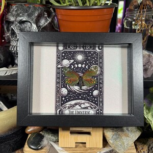 May include: A framed print with a detailed butterfly illustration and celestial elements. The artwork is set against a dark background with the words "THE UNIVERSE" at the bottom. The black frame surrounds the print, which is displayed on a cream-colored background.