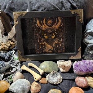 May include: A black framed art piece with gold corner accents, featuring a detailed gold illustration of a celestial design. The image is surrounded by feathers, stones, bones, and a skull, creating a mystical and gothic aesthetic.