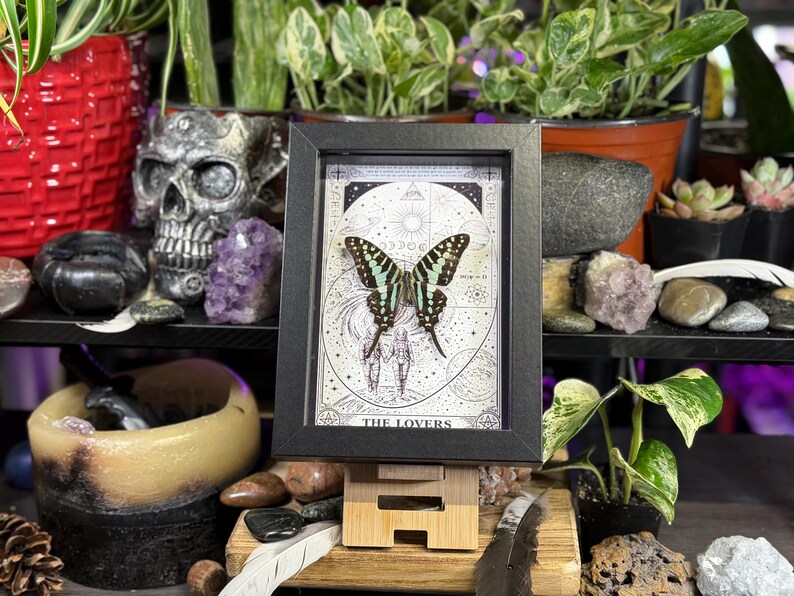 Puede incluir: Una impresi&oacute;n enmarcada que presenta una mariposa con alas negras y turquesas, sobre un fondo celestial. La impresi&oacute;n est&aacute; etiquetada como "THE LOVERS" y se muestra en un peque&ntilde;o caballete de madera. El marco es negro.