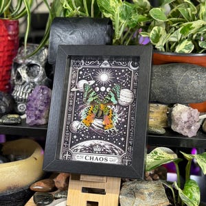 May include: A framed print with a butterfly design, set against a starry sky with planets and suns. The artwork is in a black frame and includes the word "CHAOS". The print is displayed on a wooden easel. The image has a mystical theme.