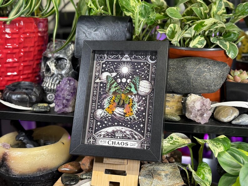 May include: A framed print with a butterfly design, set against a starry sky with planets and suns. The artwork is in a black frame and includes the word "CHAOS". The print is displayed on a wooden easel. The image has a mystical theme.