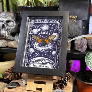 K&ouml;nnte beinhalten: Ein gerahmter Druck mit einem Himmelsdesign und einer Nachtfalter-Illustration. Das Kunstwerk ist &uuml;berwiegend blau und wei&szlig;, mit Planeten, Monden und Sternen. Der Text "The Universe" befindet sich unten. Der Rahmen ist schwarz.