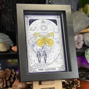K&ouml;nnte beinhalten: Gerahmtes Kunstwerk mit einem gelben Schmetterling &uuml;ber einer blau-wei&szlig;en Illustration von zwei Figuren und Himmelsk&ouml;rpern. Das Kunstwerk ist in einem dunkelgrauen Rahmen gerahmt und tr&auml;gt die Aufschrift "THE LOVERS".
