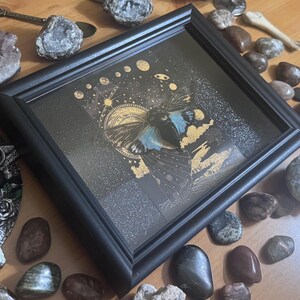 May include: A framed artwork with a celestial butterfly design. The black framed artwork has a dark background with gold and blue accents. The butterfly has blue wings and is surrounded by stars and planets. The artwork is displayed with decorative stones.