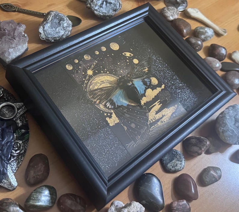 May include: A framed artwork with a celestial butterfly design. The black framed artwork has a dark background with gold and blue accents. The butterfly has blue wings and is surrounded by stars and planets. The artwork is displayed with decorative stones.