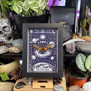 K&ouml;nnte beinhalten: Ein gerahmter Druck der Tarotkarte "Das Universum". Das Kunstwerk zeigt eine Motte mit orangefarbenen Fl&uuml;geln, Planeten, Monde und Sterne vor einem dunkelblauen Hintergrund. Der Rahmen ist schwarz und der Druck wird auf einem Holzst&auml;nder pr&auml;sentiert.
