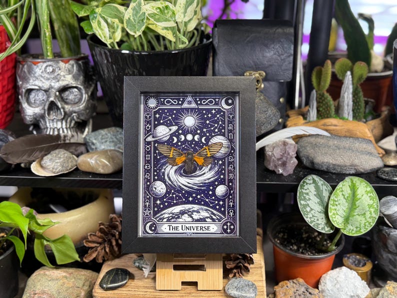 K&ouml;nnte beinhalten: Ein gerahmter Druck der Tarotkarte "Das Universum". Das Kunstwerk zeigt eine Motte mit orangefarbenen Fl&uuml;geln, Planeten, Monde und Sterne vor einem dunkelblauen Hintergrund. Der Rahmen ist schwarz und der Druck wird auf einem Holzst&auml;nder pr&auml;sentiert.