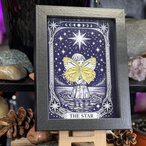 May include: Framed artwork with a celestial theme, depicting a figure with a yellow butterfly against a dark blue starry backdrop. The black frame surrounds the image, which includes the text "THE STAR".