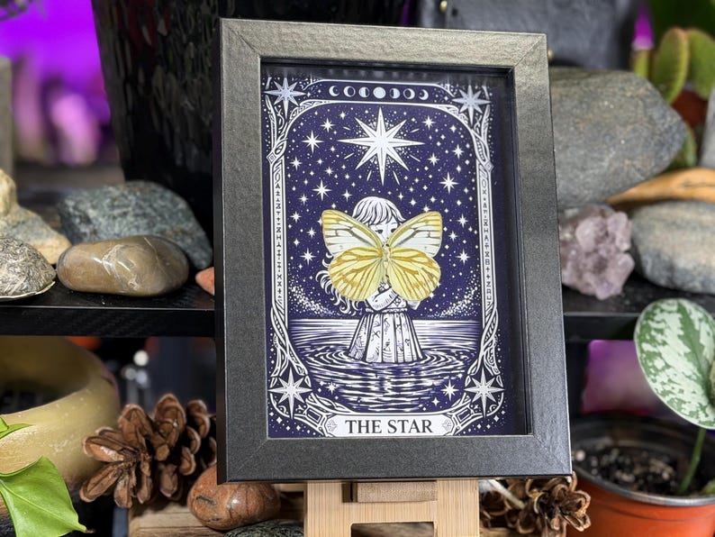 May include: Framed artwork with a celestial theme, depicting a figure with a yellow butterfly against a dark blue starry backdrop. The black frame surrounds the image, which includes the text "THE STAR".