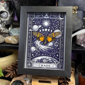 May include: A framed artwork featuring a celestial design with a butterfly. The artwork has a dark blue background with white stars, planets, and a sun. The butterfly has orange and black wings. The word "CHAOS" is at the bottom.