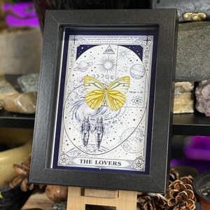 K&ouml;nnte beinhalten: Gerahmter Kunstdruck mit einem gelben Schmetterling und Himmelselementen. Das Kunstwerk mit dem Titel "The Lovers" zeigt zwei Figuren in Raumanz&uuml;gen und astrologische Symbole. Der Druck ist in einem schwarzen Rahmen, der dem mystischen Design eine elegante Note verleiht.