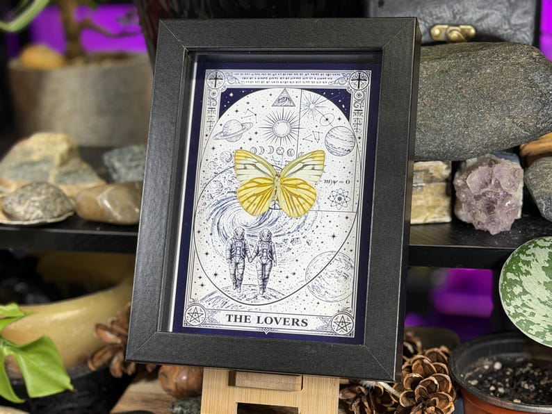 K&ouml;nnte beinhalten: Gerahmter Kunstdruck mit einem gelben Schmetterling und Himmelselementen. Das Kunstwerk mit dem Titel "The Lovers" zeigt zwei Figuren in Raumanz&uuml;gen und astrologische Symbole. Der Druck ist in einem schwarzen Rahmen, der dem mystischen Design eine elegante Note verleiht.