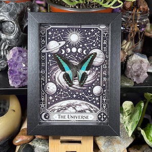 Op de afbeelding: Een ingelijste print met een vlinder met turquoise en zwarte vleugels, tegen een zwart-witte kosmische achtergrond. Het ontwerp bevat hemellichamen, sterrenbeelden en de tekst "The Universe". De lijst is zwart.