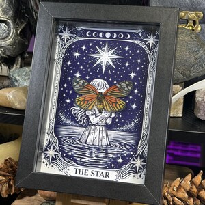 May include: A framed artwork featuring a detailed illustration of a woman holding a butterfly, set against a starry night sky. The artwork is labeled "THE STAR" and is framed in a black rectangular frame. The butterfly has orange and black wings.