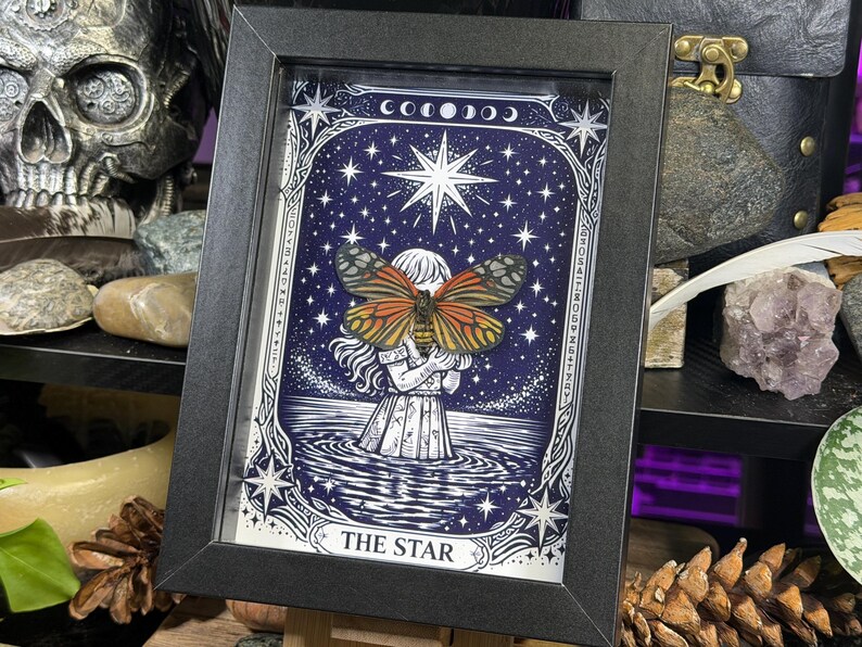 May include: A framed artwork featuring a detailed illustration of a woman holding a butterfly, set against a starry night sky. The artwork is labeled "THE STAR" and is framed in a black rectangular frame. The butterfly has orange and black wings.