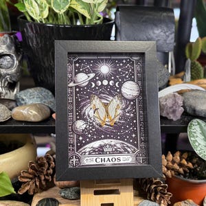 May include: A framed print with a black border, depicting a celestial design with planets, stars, and a butterfly. The print has a dark background with the word "CHAOS" at the bottom. The frame is propped up on a wooden stand.