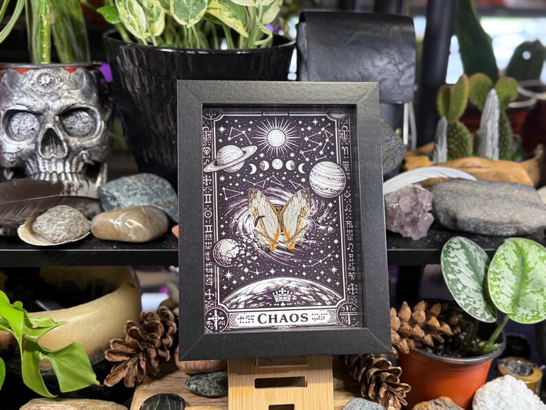 May include: A framed print with a black border, depicting a celestial design with planets, stars, and a butterfly. The print has a dark background with the word "CHAOS" at the bottom. The frame is propped up on a wooden stand.