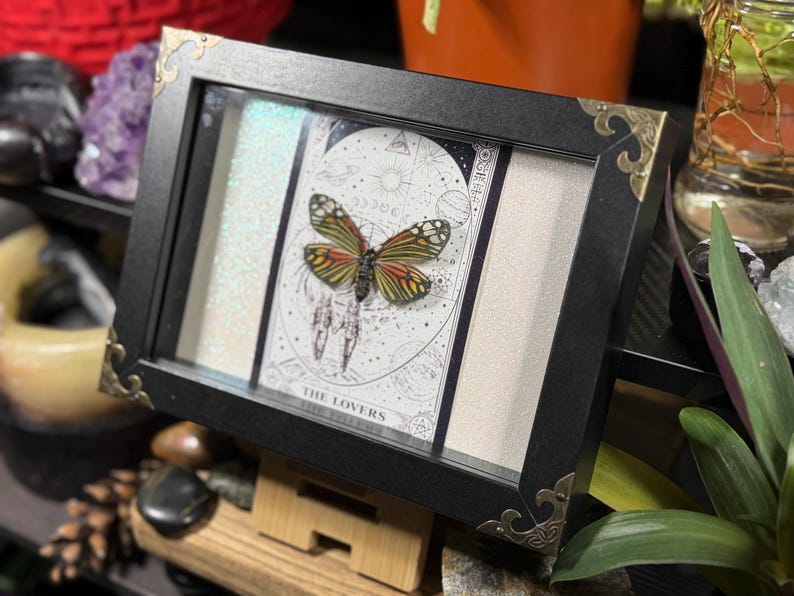 May include: A framed artwork with a butterfly and a tarot card. The black frame has decorative gold corners. The tarot card reads "THE LOVERS" and has a detailed illustration. The butterfly has orange, yellow, and black wings.