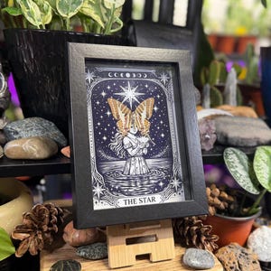 May include: A framed print of "The Star" tarot card. The image shows a figure with butterfly wings against a starry background. The black frame is displayed on a wooden stand, surrounded by plants and decorative items, creating a mystical aesthetic.