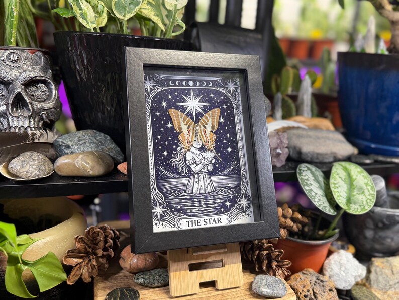 May include: A framed print of "The Star" tarot card. The image shows a figure with butterfly wings against a starry background. The black frame is displayed on a wooden stand, surrounded by plants and decorative items, creating a mystical aesthetic.