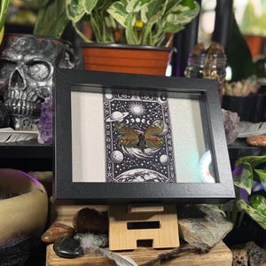 May include: A framed artwork with a black frame, displaying a detailed black and white illustration of a butterfly with celestial elements. The butterfly has orange, yellow, and green wings. The artwork is on a wooden stand.