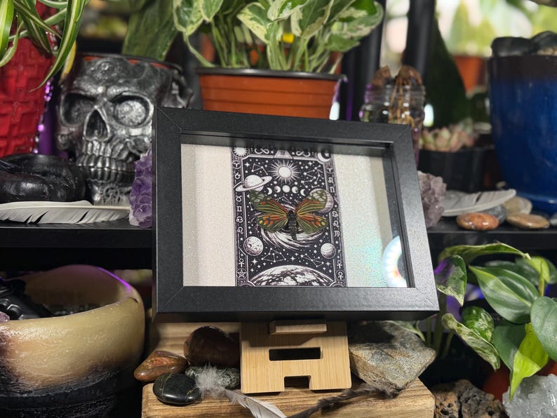 May include: A framed artwork with a black frame, displaying a detailed black and white illustration of a butterfly with celestial elements. The butterfly has orange, yellow, and green wings. The artwork is on a wooden stand.