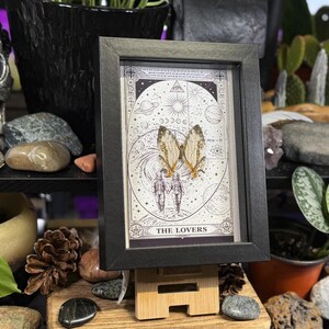 May include: A framed print featuring a tarot card design with the words "THE LOVERS" at the bottom. The artwork includes a celestial background, two figures holding hands, and butterfly wings. The frame is black.