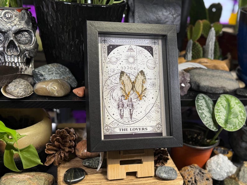 May include: A framed print featuring a tarot card design with the words "THE LOVERS" at the bottom. The artwork includes a celestial background, two figures holding hands, and butterfly wings. The frame is black.