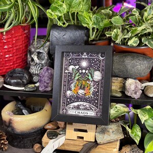 May include: A framed art print with a butterfly design in orange and yellow, set against a celestial backdrop with moons and stars. The word "CHAOS" is at the bottom. The print is displayed on a wooden stand, surrounded by plants and decorative items.