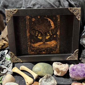 May include: A black framed art piece with gold corner accents, featuring a detailed illustration of a tree with a moth-like design, a crescent moon, and stars. The artwork is surrounded by various crystals, stones, and bones.
