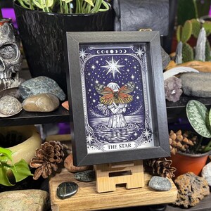 May include: Framed artwork of "The Star" tarot card. The image features a woman with butterfly wings, set against a starry night sky. The artwork is in a black frame and displayed on a wooden easel.