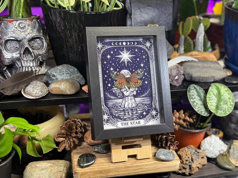 May include: Framed artwork of "The Star" tarot card. The image features a woman with butterfly wings, set against a starry night sky. The artwork is in a black frame and displayed on a wooden easel.
