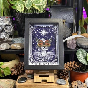 May include: A framed print of a tarot card, featuring a woman with a butterfly, standing in water under a starry sky. The print is in a black frame and displayed on a small wooden easel. The text "THE STAR" is at the bottom.