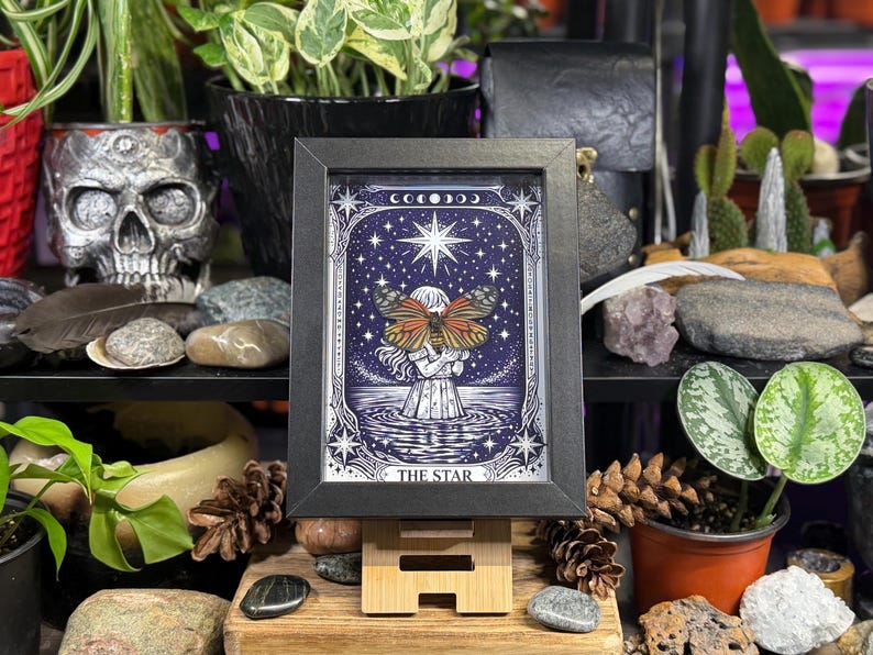 May include: A framed print of a tarot card, featuring a woman with a butterfly, standing in water under a starry sky. The print is in a black frame and displayed on a small wooden easel. The text "THE STAR" is at the bottom.