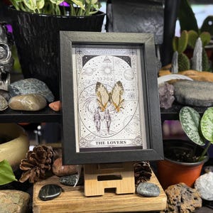 May include: A framed print of "The Lovers" tarot card, featuring a celestial design with butterflies and figures. The black frame sits on a small wooden easel, surrounded by plants, rocks, and decorative items. The overall aesthetic is mystical and bohemian.