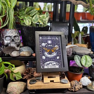 K&ouml;nnte beinhalten: Ein gerahmter Druck der Tarotkarte "Das Universum" mit einem Himmelsdesign mit Planeten, Monden und einem Schmetterling. Das Kunstwerk wird auf einer Holzstaffelei ausgestellt, umgeben von Topfpflanzen, einem Totenkopf und dekorativen Steinen. Der Rahmen ist schwarz.