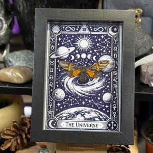 K&ouml;nnte beinhalten: Gerahmter Kunstdruck mit einem Himmelsmotiv und einer Illustration einer Motte. Das Kunstwerk zeigt einen dunkelblauen Hintergrund mit wei&szlig;en Sternen, Planeten und dem Text "The Universe". Der Rahmen ist schwarz.
