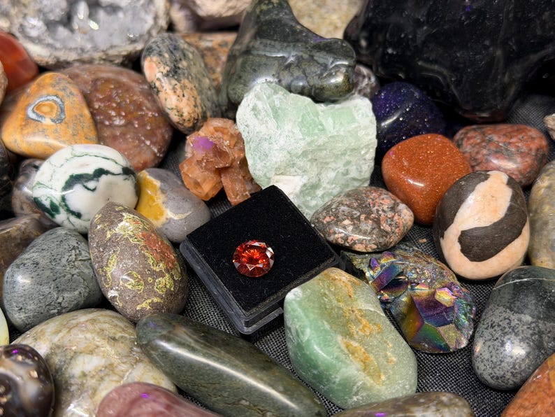 Puede incluir: Una colecci&oacute;n de piedras preciosas y cristales pulidos de diversas formas, tama&ntilde;os y colores. El surtido incluye una gema roja facetada en una vitrina negra, junto con piedras verdes, marrones y moteadas. La disposici&oacute;n es una mezcla de piedras naturales y talladas.
