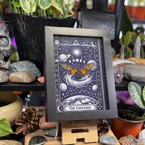 K&ouml;nnte beinhalten: Ein gerahmter Druck mit einem Himmelsmotiv, der einen Schmetterling, Planeten und den Text "The Universe" zeigt. Das Kunstwerk hat einen dunkelblauen Hintergrund mit wei&szlig;en Sternen und einem Halbmond. Der Rahmen ist schwarz.