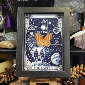 May include: Black framed artwork depicting a celestial scene with planets, stars, and the sun. The image includes two figures in space suits holding hands, a butterfly, and the words "THE LOVERS".