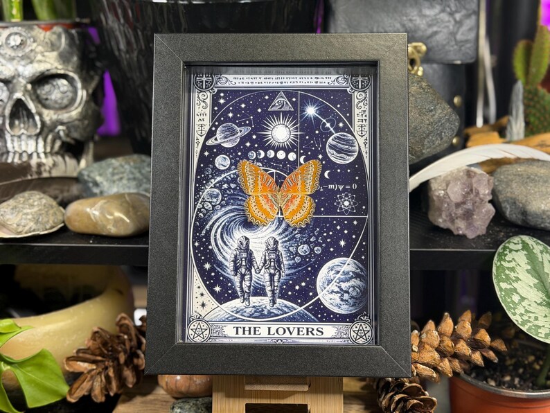 May include: Black framed artwork depicting a celestial scene with planets, stars, and the sun. The image includes two figures in space suits holding hands, a butterfly, and the words "THE LOVERS".