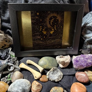 May include: A black framed art piece with a gold and black celestial design, surrounded by various crystals, stones, and bone fragments. A skull is visible in the background, along with feathers and other decorative items.