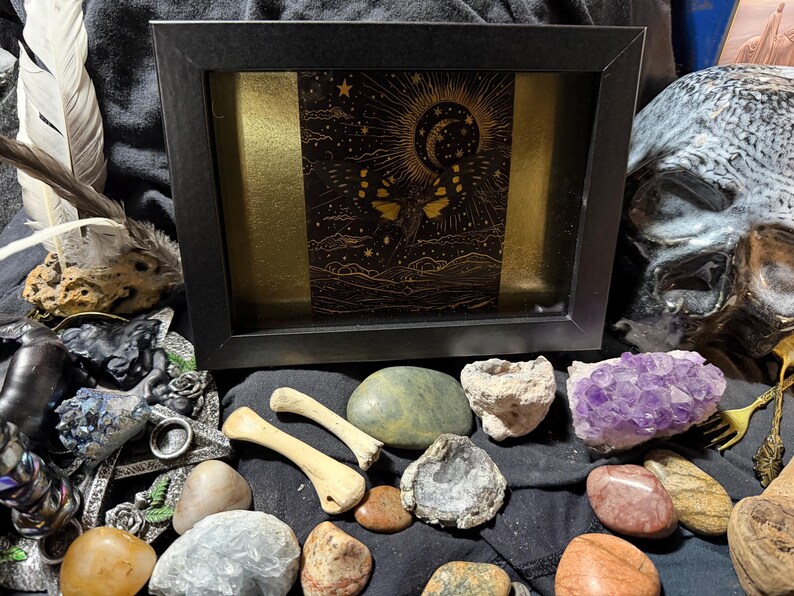 May include: A black framed art piece with a gold and black celestial design, surrounded by various crystals, stones, and bone fragments. A skull is visible in the background, along with feathers and other decorative items.